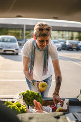One woman mature caucasian female stand at the back trunk of her car on the parking lot of the supermarket shopping mall or grocery store with vegetables food in box putting them in the vehicle