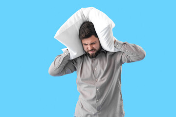 Young man with pillow suffering from loud noise on blue background