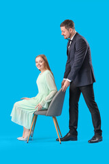 Young man pulling up chair for his wife on blue background