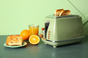 Crispy bread slices in modern toaster, cup of juice, orange and funny toast on green wooden table