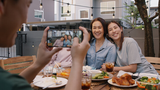 Young adult asia people hug cuddle kiss love care for mom taking photo selfie video on mobile phone camera at home picnic dining fun night party dine table. Relax older mum smile enjoy warm time meal.