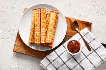 Plate with delicious wafer rolls and bowl of boiled condensed milk on white grunge background