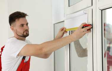 Worker in uniform using tape measure while installing roller window blind indoors, focus on hands