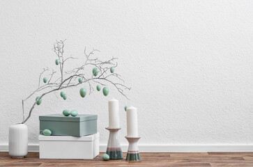Vase with tree branch, Easter eggs, candles and boxes near light wall