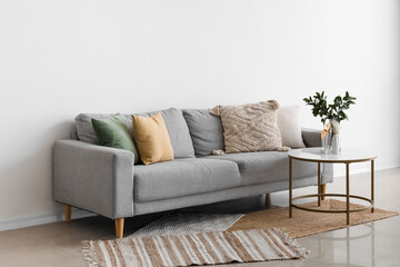 Cozy grey sofa with cushions and plant branches in vase on table near light wall