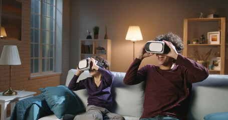 Fototapeta na wymiar Funny south asian siblings or father and son with curly hair are trying virtual reality headsets, having fun playing video games - modern technologies, family time concept 