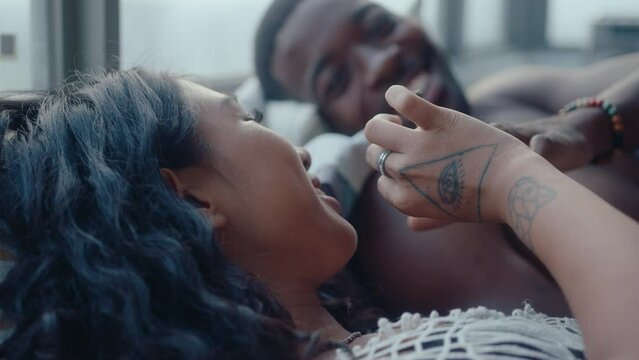 Shirtless black man lying on bed, smoking a cigarette together with girlfriend, chatting and cleaning ash from her shoulder carefully when having romantic time together at home