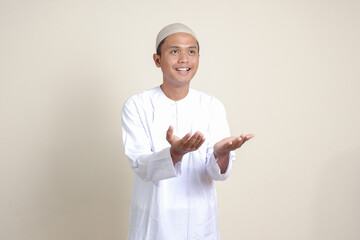 Portrait of attractive Asian muslim man in white shirt showing product and pointing with his hand and finger to the side. Isolated image on gray background