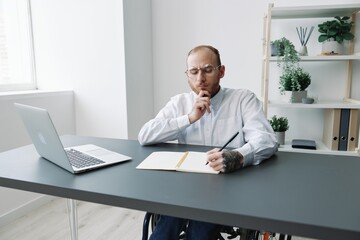 Obraz premium A man in a wheelchair, a businessman in the office works at a laptop, writing down a plan in a notebook, thoughtfulness, integration into society, the concept of working a person with disabilities