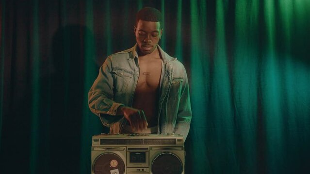 Handsome black man posing shirtless in denim jacket putting compact audio cassette in ghetto blaster and playing music in a studio with warm light and green curtain background