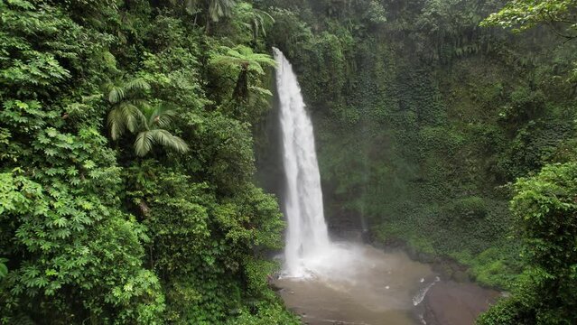 Beautiful big waterfall in green canyon, camera fly towards rushing water. Powerful stream splash down in pond, white spray fly around. Natural landmark of central Bali, one of biggest waterfalls
