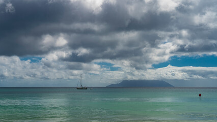 Obraz premium The smooth surface of the turquoise ocean glitters. The silhouette of the yacht is visible. The island on the horizon is hiding in the clouds. Blue sky. Seychelles. Mahe