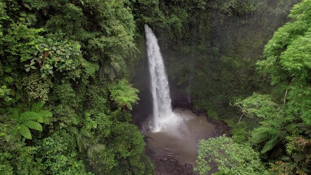 Large waterfall at deep canyon, green tropical plants around, camera fly down while looking at powerful stream of water. Nature landmark and tourist attraction of Badung Regency, Nungnung waterfall