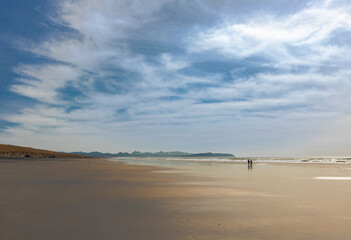Wide angle view of distant silhouetted couple on a sandy beach with dramatic wispy cloud blue sky on a sunny afternoon in the Pacific Northwest.