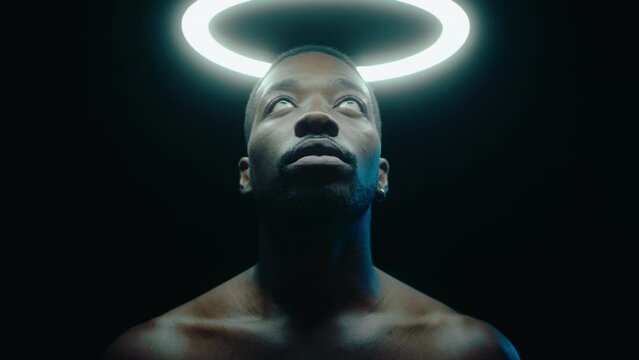 Young shirtless black man looking upwards at the ring light, posing with nimbus above the head in a dark studio. Zoom Shot