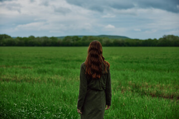 a woman with beautiful, long, red hair stands with her back to the camera in a green field in rainy, spring weather in a long raincoat