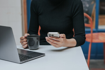 Young Asian woman using smartphone and laptop working remotely at coffee shop.