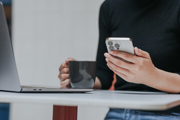 Young Asian woman using smartphone and laptop working remotely at coffee shop.