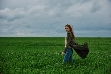 A woman stands in a green field in a raincoat and looks at the camera. Strong wind, flying hair, freedom and harmony with nature.