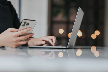 Young Asian woman using smartphone and laptop working remotely at coffee shop.