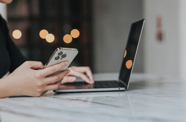 Young Asian woman using smartphone and laptop working remotely at coffee shop.