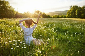 a red-haired woman in a light dress sits with her back to the camera in a field of daisies...
