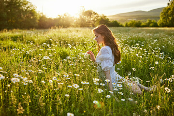 a beautiful woman in a light dress sits in a field of daisies against the backdrop of the setting...