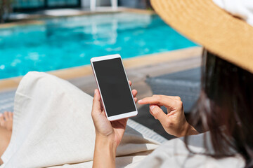 Asian woman sitting on sunbed near swimming pool using mobile phone. Young traveler girl working on cell phone during her summer holiday. Technology and lifestyle concept. Free space