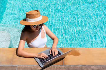 Asian woman in swimsuit using laptop working remotely in swimming pool. Young Asian traveler woman working on computer during her summer holiday. Work from anywhere, Technology and lifestyle concept