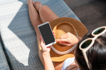 Asian woman sitting on sunbed near swimming pool using mobile phone. Young traveler female working on cellphone during her summer vacation trip. Technology and lifestyle concept. Copy space, top view