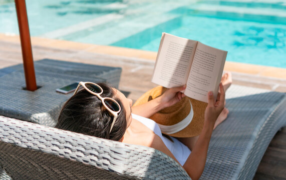 Happy girl lying down on sunbed while reading a book near Swimming Pool on travel holidays vacation, wellness Lifestyle concept. Young Asian woman relaxing at hotel on weekend. Copy space