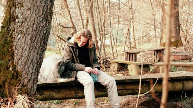 the master caresses his best friend in love. A blond young man and a gray, fluffier-than-average wolf Spitz sit together on a small bridge in an autumn landscape.