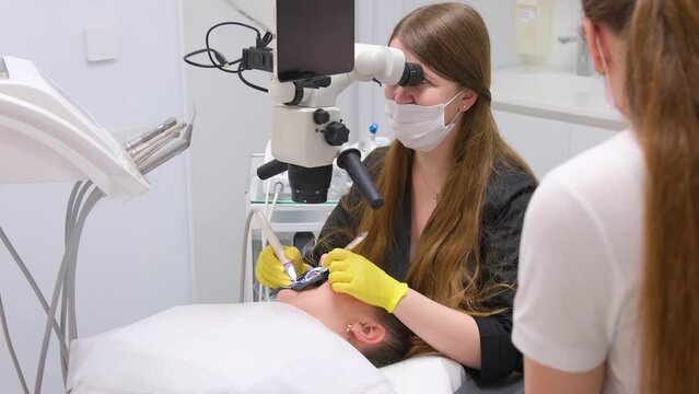 a dentist in yellow gloves in black clothes a woman works with patients a sister helps in blue gloves the girls have long loose hair Dentist operation patient through microscope 