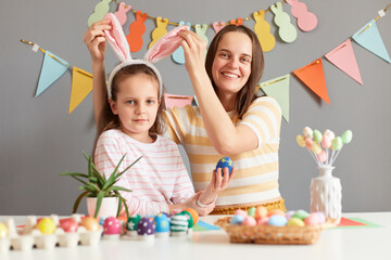 Portrait of cheerful people having fun during spring holiday, mother and daughter, woman touching kid's bunny ears painting eggs together preparing for Easter against gray decorated wall.