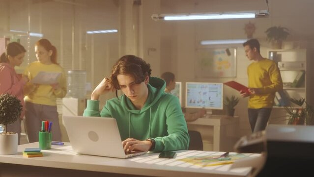 A young man is talking using a headset and typing on a laptop keyboard while sitting at a table in the office. In the background, a young creative team of managers in bright sweatshirts work.