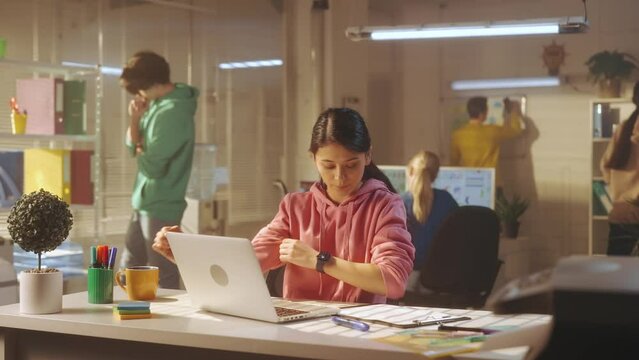 A young woman late for work runs into the office, sits at the table and starts typing on the laptop keyboard. In the background, a young creative team of managers in bright sweatshirts work.