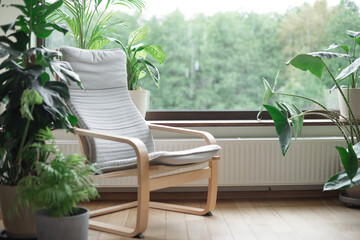Grey armchair near big panoramic window, indoor plants, monstera, palm trees. Urban jungle apartment. Biophilia design. Cozy tropical home garden. Eco friendly decor of living room.