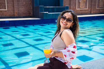 Young beautiful indian woman sitting on the pool edge with feet in water and holding glass of orange juice. Brunette girl relaxing outdoors by swimming pool. Summer holiday and vacation concept.