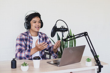 Young asian man podcaster explaining information using laptop and microphone. Technology of on-air online in broadcasting at home studio.