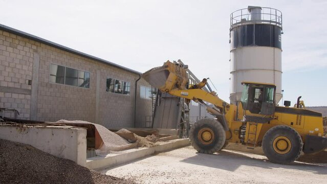 At a cement factory, a yellow industrial crane unloads sand and gravel with the trailer in front. Behind the concrete mixer where the cement is mixed. The working process of the cement factory