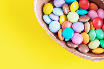 Broken chocolate Easter egg with colorful candies on yellow background