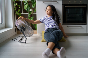 Overheating in homes. Tired overheated young Asian woman sitting on floor in kitchen near electric...