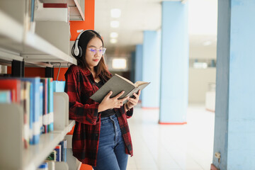 Smart young Asian student in casual style lean on library bookshelf and reading a book while listening to the music. 