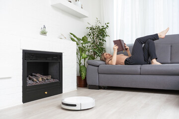 Modern robot vacuum cleaner near sofa with resting woman in room