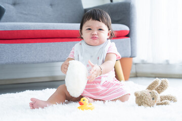 Happy cute little 7 months old multiracial, asian and caucasian, newborn baby girl sitting on floor in living room at home, holding pillow, playing alone with yellow duck toy and doll