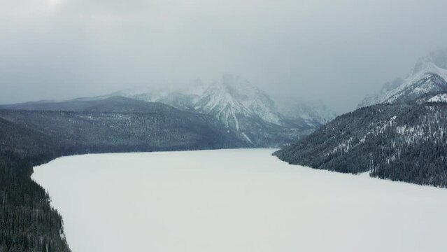 Aerial moving over frozen lake with large mountains covered in clouds behind