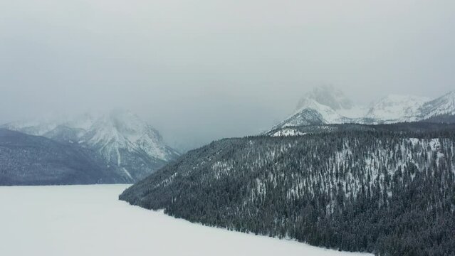 Aerial moving over frozen lake and forest towards large mountains in fog