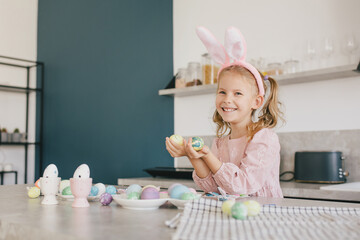 Happy cute little girl decorating Easter eggs using brush and paint on the kitchen.