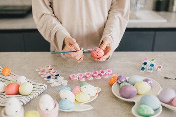 Young woman decorating Easter eggs using brush and paint on the kitchen.