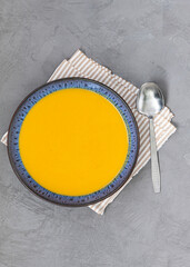 Fototapeta na wymiar Pumpkin soup in a ceramic plate with a spoon on a striped cloth napkin. Gray texture background. Top view
